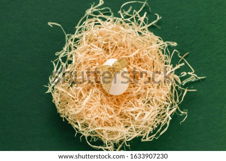 White egg with a golden bow in a hay nest and sweets around on the green background. Holy flatlay. Copyspace for the wording. Easter holidays concept