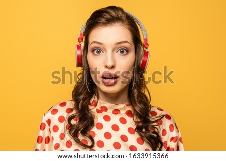 shocked young woman in wireless headphones looking at camera isolated on yellow
