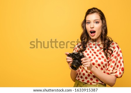 shocked young woman holding digital camera while looking at camera isolated on yellow