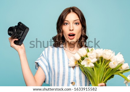 surprised young woman holding digital camera and bouquet of white tulips isolated on blue