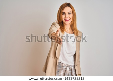Redhead caucasian business woman standing over isolated background smiling friendly offering handshake as greeting and welcoming. Successful business.
