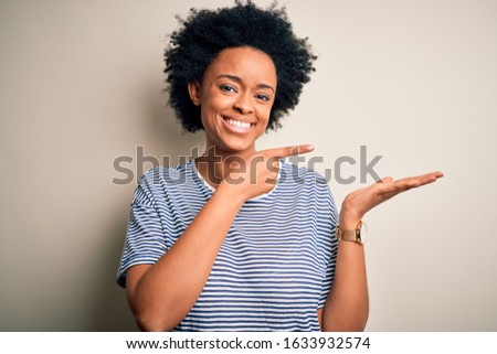 Young beautiful African American afro woman with curly hair wearing striped t-shirt amazed and smiling to the camera while presenting with hand and pointing with finger.