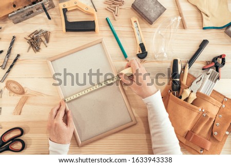 Female carpenter tape measuring picture frame in small business woodwork workshop, top view of hands