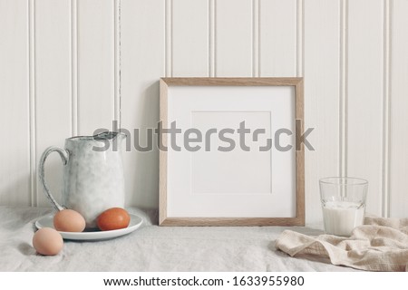 Spring breakfast still life scene. Square empty wooden frame mockup with chicken eggs,, glass of milk and ceramic jug. Easter food and drink concept. Farmhouse, Scandinavian design.