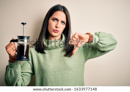 Young woman with blue eyes making cafe using coffe maker standing over white background with angry face, negative sign showing dislike with thumbs down, rejection concept