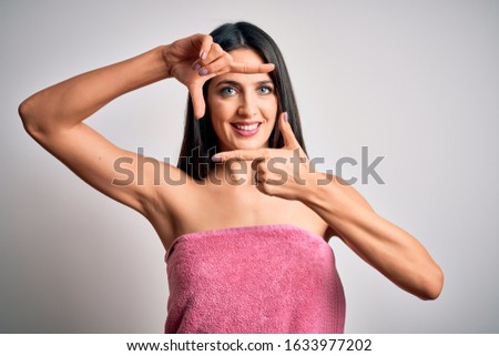 Young brunette woman with blue eyes wearing shower towel after bath over white background smiling making frame with hands and fingers with happy face. Creativity and photography concept.
