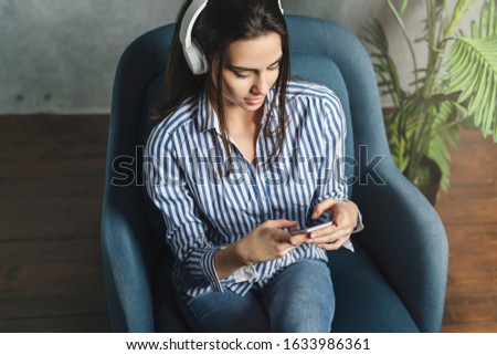 Girl in earphones listening to music in online app sitting on sofa in her own apartment. Young lady downloading audio apps, digital tools and solutions on smartphone. Streaming from social networks.