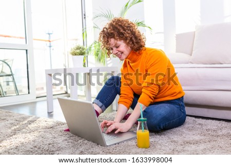 Image of a young red hair woman drinking orange juice and surfing in the internet at home. Freelancer female working on laptop from home