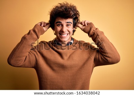 Young handsome man wearing casual shirt and sweater over isolated yellow background Smiling pulling ears with fingers, funny gesture. Audition problem