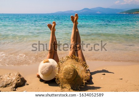 mom and daughter sunbathe on a sandy beach in hats, legs up, blue sky and blue sea