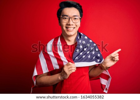 Young chinese patriotic man wearing united states of america flag over red background very happy pointing with hand and finger to the side
