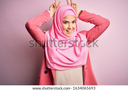 Young beautiful girl wearing muslim hijab standing over isolated pink background Posing funny and crazy with fingers on head as bunny ears, smiling cheerful