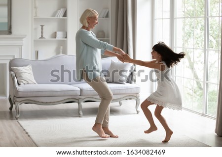 Happy middle-aged grandmother play dancing and swirling in living room with cute little granddaughter, energetic senior granny have fun engaged in childish funny activity with small preschooler child Royalty-Free Stock Photo #1634082469