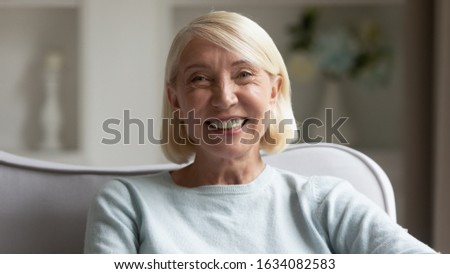 Close up headshot portrait of happy senior woman sit on couch look at camera posing show healthy white teeth, smiling middle-aged mature female feel overjoyed and pleased, optimistic elderly concept