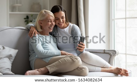 Happy elderly mother and grownup daughter sit on couch relaxing together posing for self-portrait picture on cellphone, smiling mature mom and adult millennial girl take selfie on modern smartphone