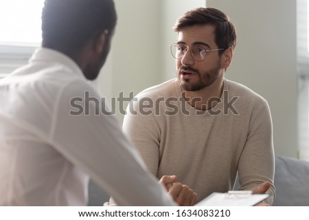 Man consulting with practitioner, African psychologist holding clipboard with card sitting in front of patient listens his mental health complaints. Job interview process applicant and HR manager Royalty-Free Stock Photo #1634083210