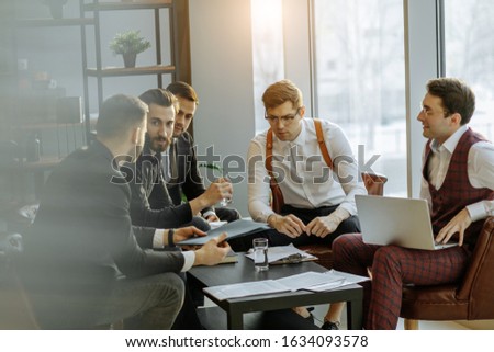 young caucasian business crew coworking, analyzing and commenting reports sitting at table together, dressed in tuxedo. isolated in modern office, big panoramic window in the background