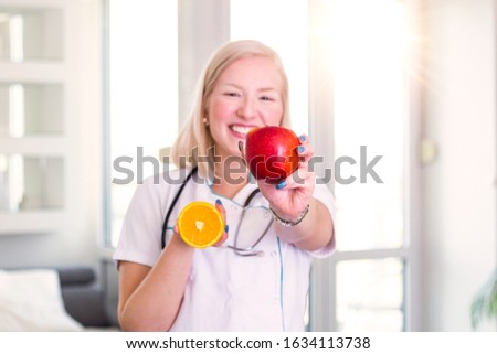 Portrait of beautiful smiling female nutritionist holding an apple and orange. Nutritionist with healthy fruit, juice and measuring tape. Dietitian working on diet plan.