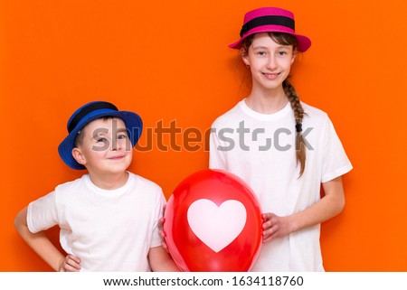 A 9 year old boy and 11 year old girl  posing with red baloon and white heart on the baloon.  Valentines day concept.Orange studio background.They enjoy being together.Copy space mock up.
