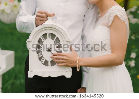 White table clock in the hands of the bride and groom. Wedding decor.