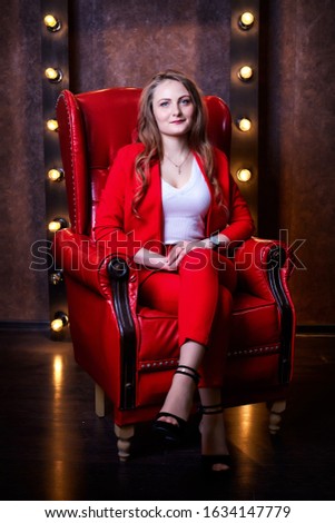 Girl in red costume sitting in red chair in the dark room. Model posing during fashion photoshoot