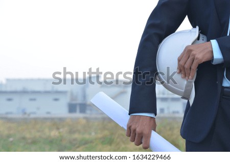 Engineer holding white helmet with blueprints for workers security on working site background.