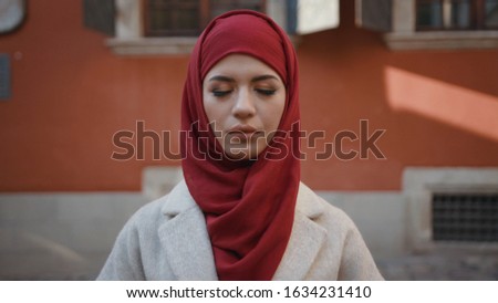 Portrait of lovely arabian young woman with closed eyes outdoors. Beautiful Muslim girl opening her eyes and smiling at camera on the street.