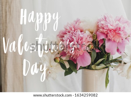 Happy Valentine's day text handwritten on lovely peony bouquet in sunny light on rustic window sill. Stylish pink and white peonies in vase on wooden background. 