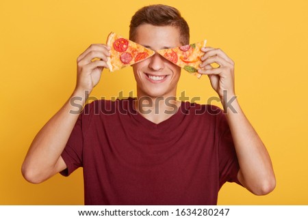 Image of cheerful handsome young boy holding two pieces of pizza in both hands, having beautiful smile, covering his eyes with pizza, being in high spirits, fastfood lover. People and food concept. Royalty-Free Stock Photo #1634280247
