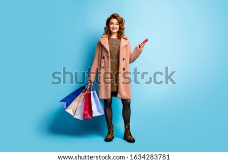 Full size photo of positive feminine lady tourist walk fun use smartphone to pay easy shopping hold many bags wear season coat dress tights  cotton boots isolated over blue color background