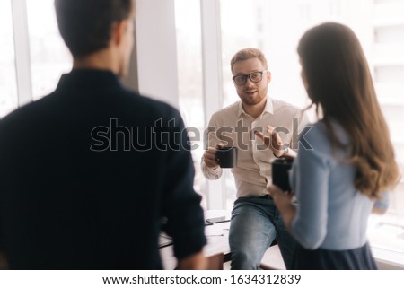 Young business team sharing ideas during coffee break. Two handsome young businessmen and lady in casual clothing are holding cups of coffee, talking and smiling, standing indoors the office building.
