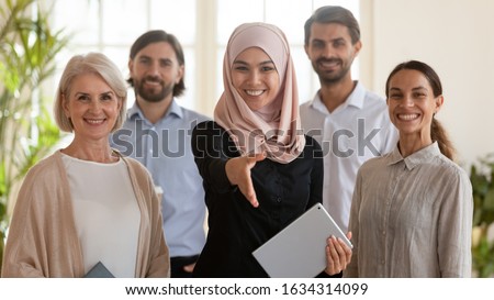 Smiling Arabian businesswoman stretch hand get acquainted greeting with client or customer pose together with diverse colleagues in office, happy multiracial employees look at camera show unity Royalty-Free Stock Photo #1634314099