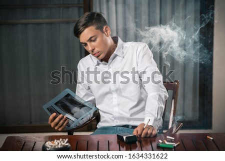 The guy is considering a picture of an X-ray of the lungs, sitting at a table with an ashtray on it, cigarettes and a lighter with a cigarette in his hand. Awareness of the consequences of smoking.