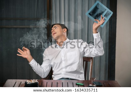 Humility of a guy posing sitting at a table on which an ashtray stands, lie cigarettes and a lighter, with a picture of fluorography and a cigarette in his hand. The consequences of smoking. 