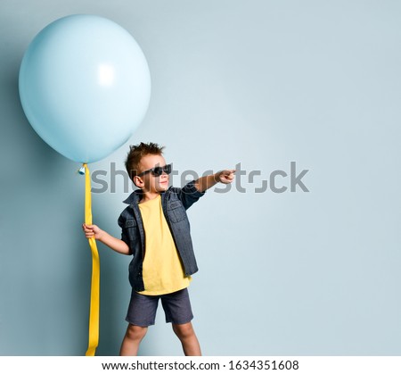 Blond boy in stylish casual clothing, sneakers and sunglasses standing near blue air balloon and pointing right over blue background. Trendy children clothes, holiday, party concept