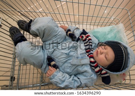 funny little boy in blue ski suit and striped scarf lies and sleeps in grocery cart from store