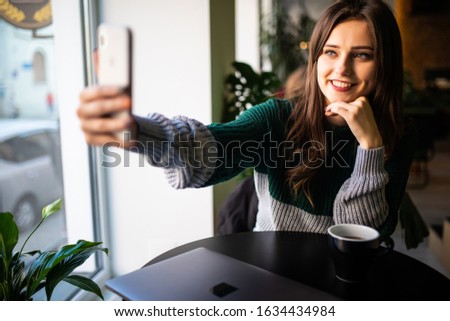Young woman in cafe sitting at table holding cup of hot coffee taking selfie pictures on phone