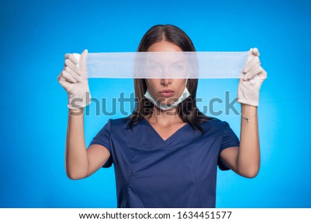 A brunette posing while standing against a blue background in a protective mask on her face, a blue surgical suit, wearing white medical gloves on her hands, with stretched medical bandages covering h