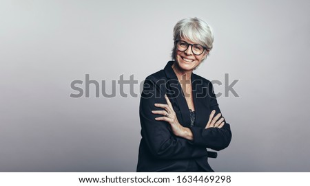 Portrait of a happy senior businesswoman looking at camera. Senior woman entrepreneur with her arms crossed standing against grey background.