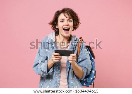 Cheerful young girl student in denim clothes backpack isolated on pastel pink background in studio. Education in high school university college concept. Mock up copy space Play game with mobile phone