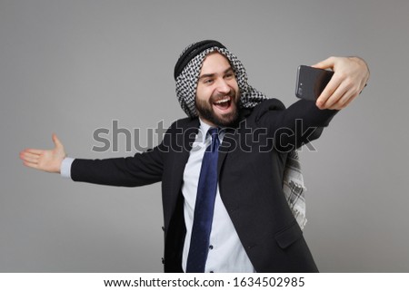 Cheerful arabian muslim businessman in keffiyeh kafiya ring igal agal suit isolated on gray background. Achievement career wealth business concept. Doing selfie shot on mobile phone spreading hands