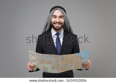 Smiling young bearded arabian muslim businessman in keffiyeh kafiya ring igal agal classic black suit shirt tie isolated on gray background. Achievement career wealth business concept. Hold city map