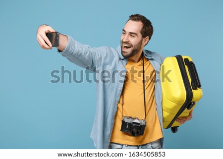 Funny traveler tourist man in summer clothes with photo camera isolated on blue background. Passenger traveling abroad on weekends. Air flight journey. Hold suitcase doing selfie shot on mobile phone