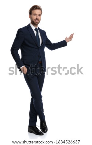 happy young businessman smiling and presenting to side, holding hands in pockets and standing isolated on white background, full body