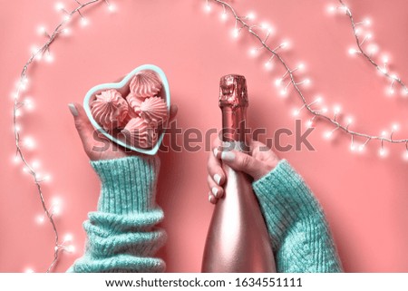 Valentine flat lay, top view on pink background. Light garland, woman hands with champagne bottle and heart shape bowl with pink marshmallow sweets. St. Valentine's day February 14 concept.