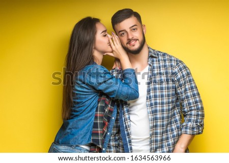 Image of brunette woman whispering secret or interesting gossip to surprised man in his ear isolated over yellow background.