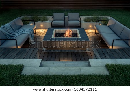 Zone for relax with a wooden floor and a tiled stair outdoors. There is a burning fire pit, gray sofas and armchairs, plaid, luminous lamps. Horizontal. Royalty-Free Stock Photo #1634572111