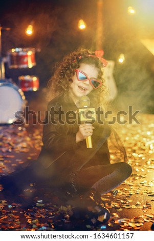Beautiful girl with curly hair wearing leather jacket, red sunglasses, boots sing into a wireless microphone for karaoke in recording studio.