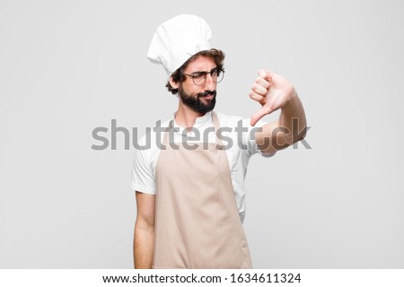 young crazy chef feeling cross, angry, annoyed, disappointed or displeased, showing thumbs down with a serious look against white wall