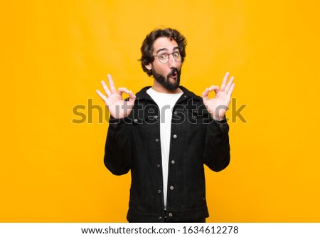 young crazy handsome man feeling shocked, amazed and surprised, showing approval making okay sign with both hands against orange wall